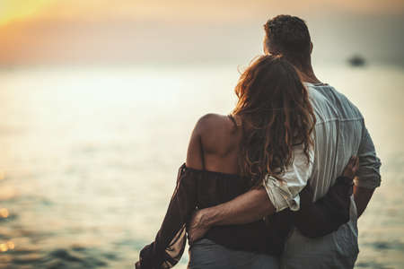 A loving couple is having fun and hugging on the empty sandy sea beach at sunset.They are looking...