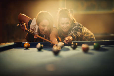 Young smiling cheerful two young women are playing billiards in bar after work. They are involved in recreational activity.の写真素材