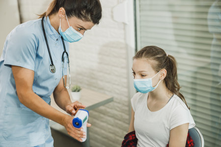 Nurse measuring temperature of a cute female teenager in waiting room during pandemic.の写真素材