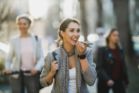 A smiling young business woman talking voice message on a smart phone on her way to work.の写真素材