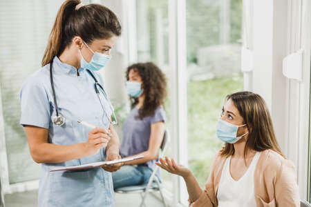 Nurse talking to young pregnant woman and taking notes before covid-19 vaccine at hospital waiting...