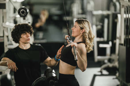 A young muscular woman is doing training at machine supporting by coach in the gym.の写真素材