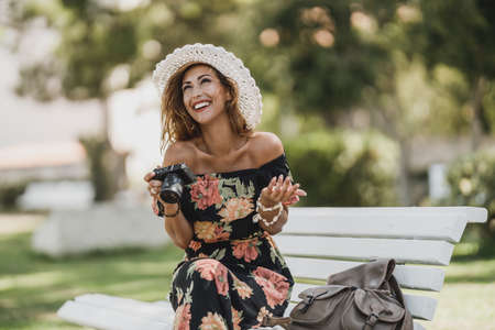 Attractive cheerful woman with digital camera sitting on a bench in the park and enjoying.