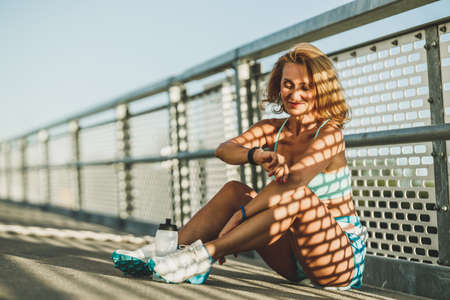 Middle age sportswoman looking on smart watch while resting after outdoor training on a bridge.の写真素材