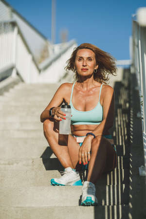 Middle age sportswoman sitting on stairs of a bridge and resting after hard training.の写真素材