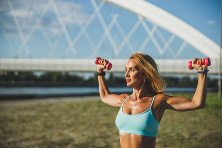 Adult fit woman sportswoman doing exercises with dumbbells during workout near the river.の写真素材