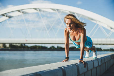 Adult woman exercising push-ups during sports training near the river in the city.の写真素材