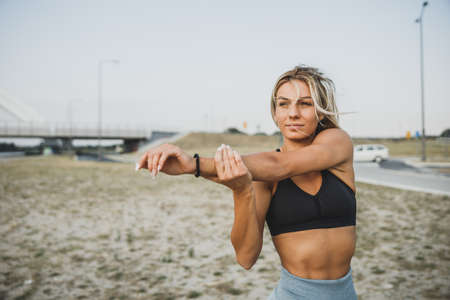 Portrait of a fit young woman warming up and stretching her arms near the river bridge.の写真素材