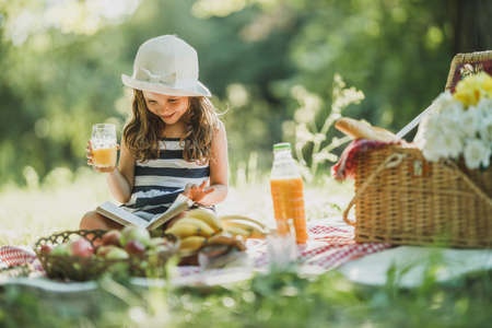 A cute smiling little girl reading a book while enjoying picnic day in nature.の写真素材