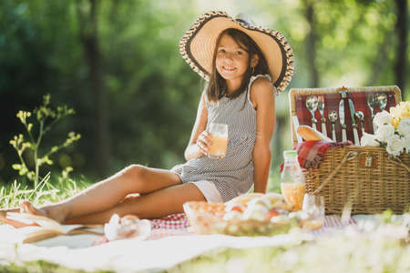 A cute smiling little girl drinking orange juice while enjoying picnic day in nature.の写真素材