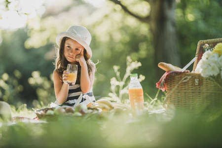 A cute little girl drinking orange juice and thinking while enjoying picnic day in nature.の写真素材