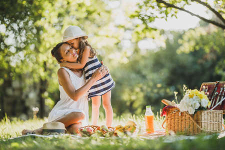 Cute little girl hugging and kissing her beautiful mom while enjoying a picnic day in nature.の写真素材