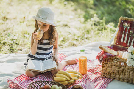 A cute smiling little girl drinking orange juice and reading a book while enjoying picnic day in nature.の写真素材