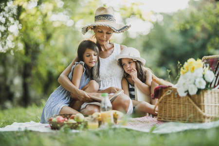 Mother and her cute daughters having a picnic. They are reading book and spending a day in nature during Covid-19 crisis.の写真素材