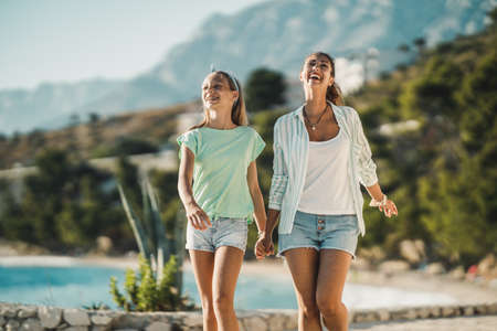 A smiling teenage girl and her young mom walking near the sea and enjoying summer vacation at the beach.の写真素材