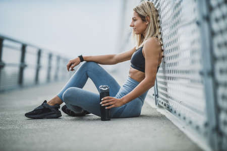 Beautiful fit woman holding a bottle of water and sitting on a bridge, in the city before training...
