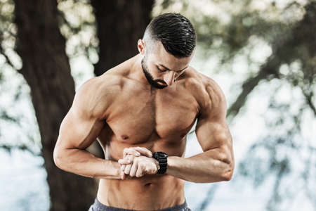 Strong bodybuilder with naked muscular torso standing in nature and looking at smartwatch ready for workout.の写真素材