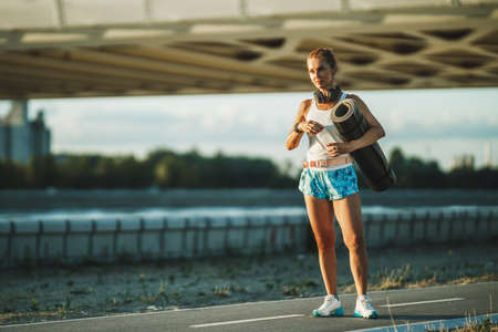 A middle age sportswoman holding a yoga mat and water bottle near to river before training in the city.の写真素材