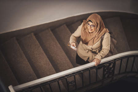 Middle aged Muslim businesswoman wearing hijab is standing on stairs of her home building and tipping messages on her smartphone.の写真素材