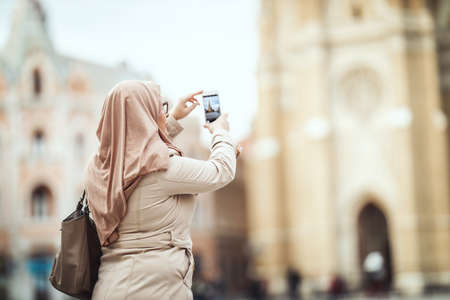 Middle aged Muslim woman wearing hijab standing in urban environment and taking photos by her smartphone.の写真素材