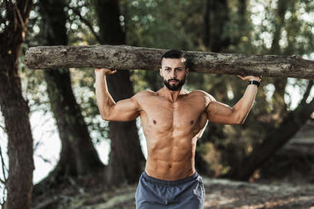 Muscular young man with naked torso doing exercises with timber in the nature, near the river.の写真素材