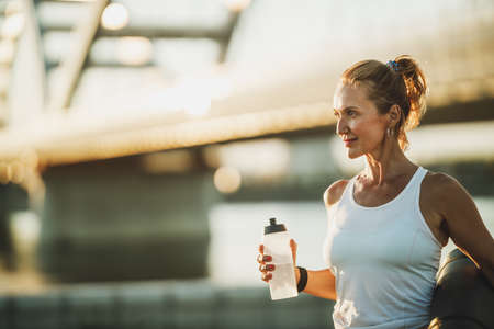 Middle age sportswoman holding water bottle before hard training near to river, in the city.の写真素材