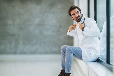 Tired doctor sitting in an empty hospital hallway and worriedly looking out the window.