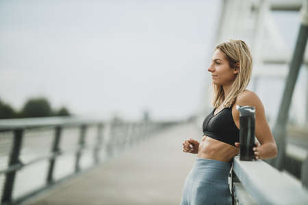 Beautiful fit woman standing and holding a bottle of water after hard training on the bridge in the city.の写真素材