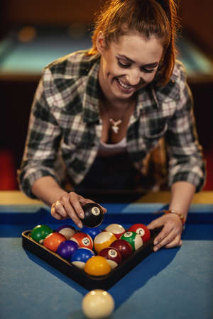 A young smiling woman places billiard balls on a pool table to play.