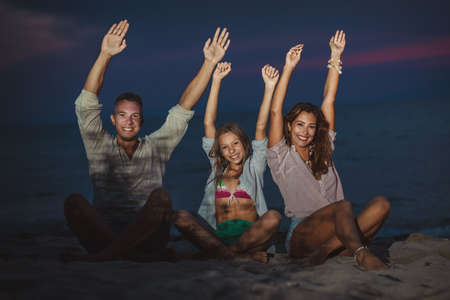 A beautiful happy family is having fun on the empty sea beach sitting and playing on the sand in the evening. They are holding hands up.の写真素材
