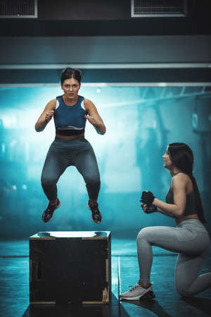 A young fit woman is doing hard exercise at the gym. She is jumping on the boxes supporting by female coach.の写真素材