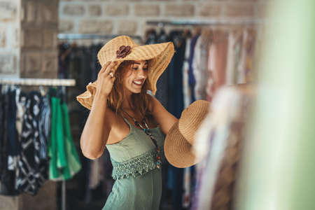 A beautiful smiling young woman is walking along the streets of a Mediterranean town and enjoys in summer sunny day.  She chooses a new hat, wanting to buy it.の写真素材
