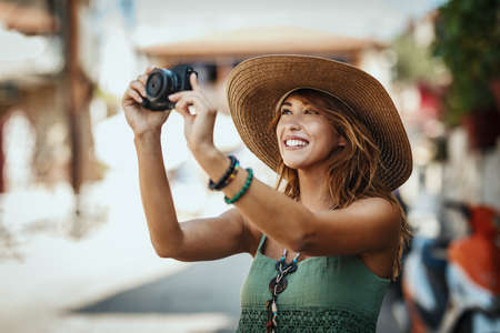 A beautiful smiling young woman with straw hat is walking around the streets of a Mediterranean town and enjoys in summer sunny day. She is making photos for memories.の写真素材