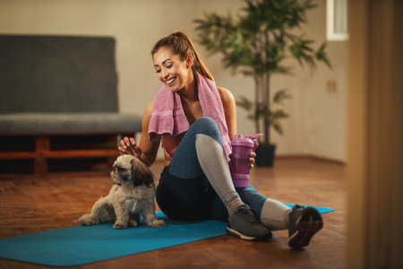 Young woman is doing stretching exercises in the living room at home supporting by her pet dog. She is resting on floor mat in morning sunshine.の写真素材