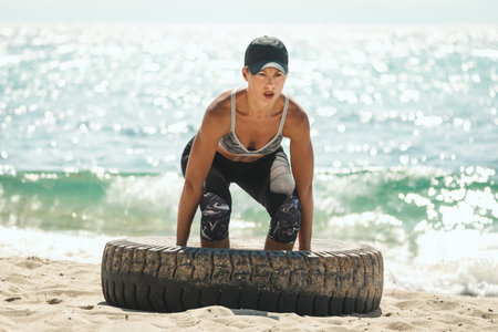 Middle age muscular woman is flipping a tire on training at the sea beach in summer sunny day.の写真素材
