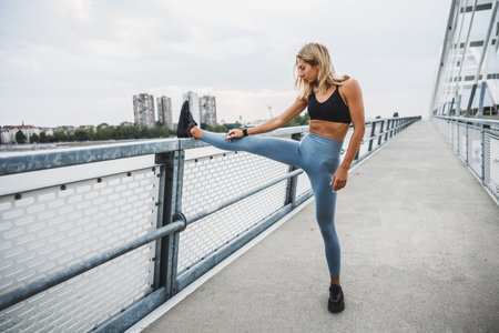 Muscular young woman warming up and stretching her legs while exercising on the bridge in the city.の写真素材