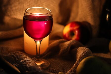 A glass full of red wine with candle muffled light shining through glass behind it and fruis on a sackcloth background.の写真素材