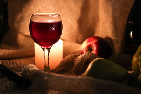 Still life. A glass of red wine and candle light behind it on table in a half light with fruits on sackcloth backgroundの写真素材