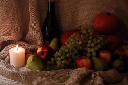 Autumn harvest still life. Group of fruits: red apples, green pears, white grapes an orange pumpkins on sackcloth backgound with bottle of wine and soft candle light.の写真素材