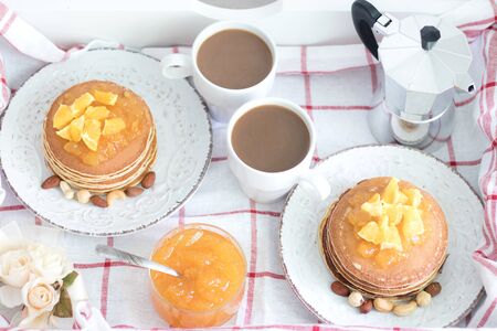 Romantic traditional breakfast for two on a tray. American pancakes with orange jam and nuts on vintage plates and 2 white coffee cups with italian metal coffee pot on a checkered cloth. Top viewの写真素材