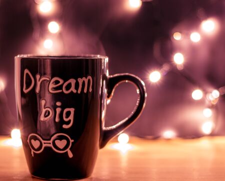 Coffee mug with letters dream big with steam on a wooden top and christmas small lights on a dark background. Copyspace. Front viewの写真素材