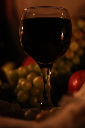 Glass of red wine with reflection of two candles flames on it close up and fruits: grapes, apple on sackcloth background. Autumn evening home warmth still life with dark colors. Happy Thanksgiving dayの写真素材