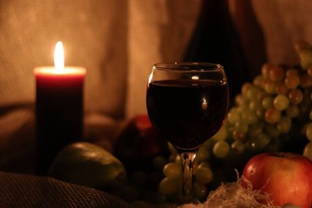 Autumn warm still life. Thanksgiving post card. Glass full of red wine with candle flame reflection on it and lit brown candle with fruits: fresh green grapes, apples and pears on sackcloth backgroundの写真素材