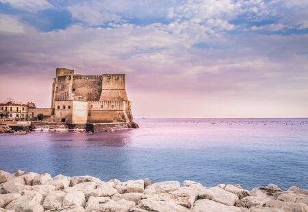 Castel dell'Ovo in Naples Italy, Campania. Picturesque view with seascape on Egg castle and Tyrhenian blue sea with white stones shore and fairy pink and violet sky of pastel colors on summer day.の写真素材