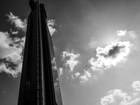 Black and white photo of single skyscraper with small plane flying near it on a clody sky background. Urban art scene with space for text.の写真素材