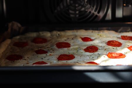Raw focaccia dough with sliced fresh tomatoes cherry and sea salt on a baking sheet and black tray in the oven ready to be baken.の写真素材