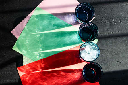 Shadow and light play with color long shades of blue glasses in a row with ice and water on a colorful pink, green and red paper on a black background. Top view, text space.の写真素材