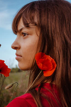A young, white teen girl portrait close-up in profile looking confidently straight with brown eyes and dark long hair with bang on nature meadow with red blooming poppies flowers bouquet near her faceの写真素材