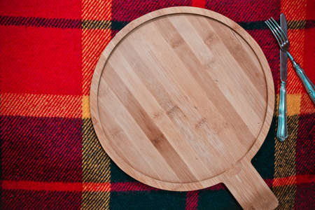 Cold season menu background, cooking or baking: round empty wooden bamboo cutting board with silver knife and fork kitchen utensils on checkered woolen red yellow plaid textile, top view copy space.の写真素材