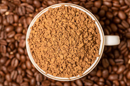 On top of the sprinkled brown beans is a white cup of ground coffee. Close-up.